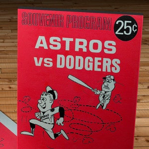 May include: A red and white souvenir program for a baseball game between the Astros and Dodgers. The program features a cartoon illustration of two baseball players, one running and the other swinging a bat. The program is priced at 25 cents and includes the text "ASTRODOME" and "Houston, Texas".
