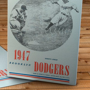 May include: A vintage 1947 Brooklyn Dodgers baseball program cover featuring a black and white illustration of a baseball game. The program cover is printed on a textured, light gray background with red and blue accents. The text "1947 Brooklyn Dodgers" is printed in red and blue on the bottom of the cover.