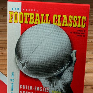 Może przedstawiać: Plakat vintage z klasycznym futbolem, z czerwonym tłem i żółtym tekstem. Plakat przedstawia dłoń trzymającą piłkę nożną, z tekstem "PHILA EAGLES VERSUS GREEN BAY PACKERS SEPT 13 1945".