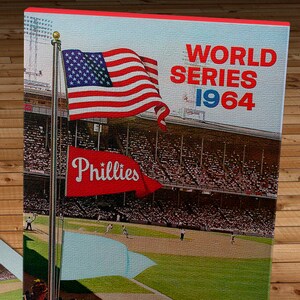 May include: A canvas print of a baseball game scene from the 1964 World Series. The American flag and a Phillies pennant fly above the stadium. The words "World Series 1964" are printed in red.