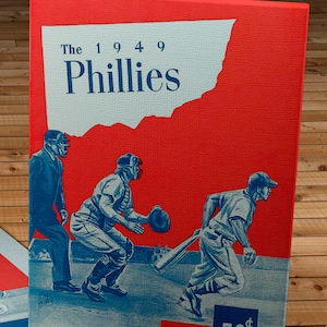 May include: A vintage 1949 Philadelphia Phillies baseball card featuring three players in action. The card is red with a white outline of the state of Pennsylvania. The card has a blue and white design with the text "The 1949 Phillies" and "50¢".