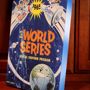 May include: A vintage 1965 World Series program cover featuring a blue and white space-themed design. The cover depicts two baseball teams, the Twins and the Dodgers, flying in rocket ships. The text "OUT OF THIS WORLD SERIES" is prominently displayed in orange and black. The program costs fifty cents.