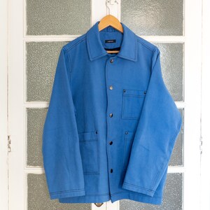 K&ouml;nnte beinhalten: Eine blaue Arbeitsjacke mit Knopfleiste und mehreren Taschen. Die Jacke hat ein schwarzes Etikett mit dem Text "LARIVIERE" auf der linken Brustseite.