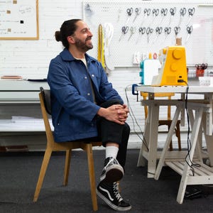 K&ouml;nnte beinhalten: Ein Mann in einer blauen Jeansjacke und schwarzen Hosen sitzt auf einem Holzstuhl. Er schaut nach rechts und l&auml;chelt. Im Hintergrund steht eine gelbe N&auml;hmaschine auf einem wei&szlig;en Tisch.