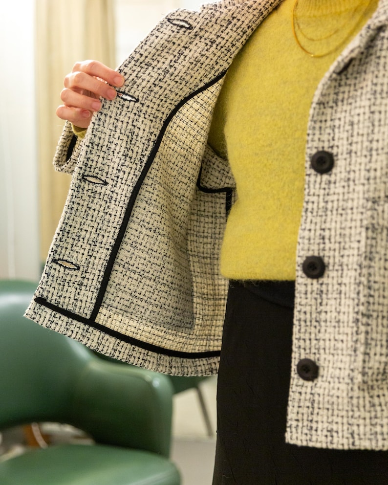 K&ouml;nnte beinhalten: Eine wei&szlig;e und schwarze Tweedjacke mit schwarzem Besatz. Die Jacke hat einen Knopfverschluss und ist offen, so dass das schwarze Futter sichtbar ist.