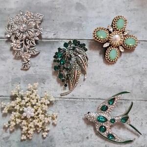 May include: A collection of decorative brooches in different styles. The brooches include a silver floral spray, a green leaf design, a gold and green flower, a pearl and crystal snowflake, and a green and silver bird.