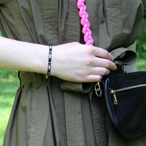 Puede incluir: Un brazalete de cuentas negras y blancas con un dije plateado en la muñeca de una persona. La persona lleva una camisa verde y una correa trenzada rosa.