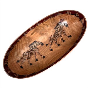 May include: An oval wooden serving bowl with a red and black geometric design around the rim. The bowl features two black and white zebra illustrations.