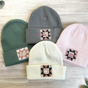 May include: Four ribbed knit beanies in shades of green, gray, pink, and white. Each beanie features a square patch with a floral and geometric design. The beanies are arranged on a light wood-grain surface.