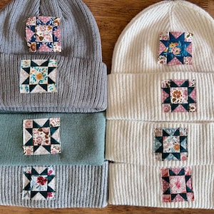 May include: A collection of ribbed knit beanies in gray, teal, and cream. Each beanie features a square patchwork design with floral and geometric patterns. The beanies are displayed on a wooden surface.