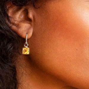 Puede incluir: Un pendiente colgante de plata con una pequeña piedra preciosa de citrino amarillo de corte cuadrado suspendida de un fino gancho de metal. El pendiente cuelga del lóbulo de la oreja, mostrando el color amarillo dorado translúcido de la piedra.