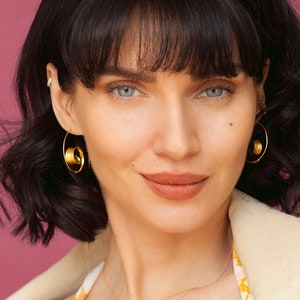 May include: A woman with dark brown hair wearing gold hoop earrings with a swirl design. She is wearing a beige blazer and a floral patterned top.