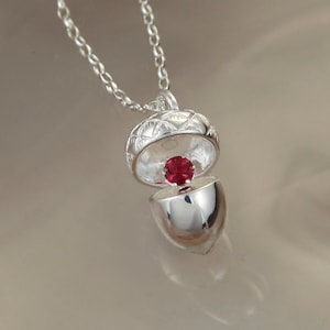 May include: A silver acorn necklace with a red gemstone inside. The acorn is open and the gemstone is visible. The necklace is on a white background.