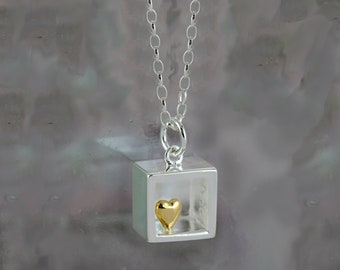 May include: A silver necklace with a square pendant featuring a small gold heart inside. The pendant is open on one side, creating a frame-like effect.
