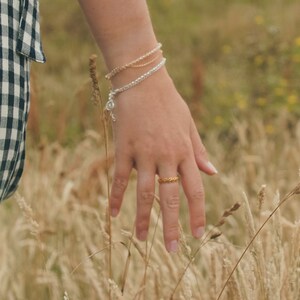 May include: A hand wearing a gold ring and a stack of beaded bracelets, reaching out towards a field of tall, dry grass.