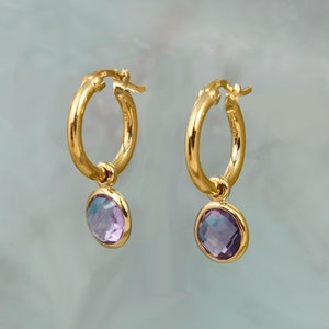 May include: A pair of gold hoop earrings with a dangling, oval-shaped, iridescent purple gemstone. The earrings have a polished finish and are set against a soft, light blue background. The jewelry is designed for pierced ears.