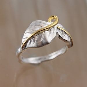 May include: A silver ring with a gold leaf design. The ring is made of two leaves, one on top of the other, with a gold band around the top leaf.
