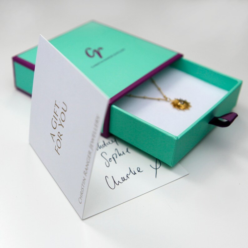 May include: A teal gift box with a purple ribbon is open, revealing a gold necklace with a small pendant. The box is sitting on a white surface. A white card with the text "A GIFT FOR YOU" is visible in the background.