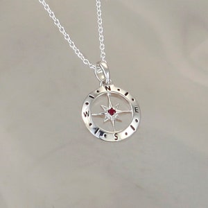 May include: Silver compass necklace with a red gemstone in the center. The compass features the letters N, S, E, and W around the edge.