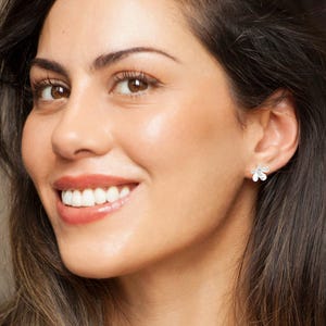 Peut inclure: Une petite boucle d'oreille à clou argentée en forme de fleur à cinq pétales. La boucle d'oreille présente une finition métallique avec un point central, portée sur le lobe de l'oreille. Le design est simple, composé de cinq pétales arrondis.