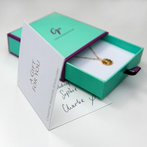 May include: A teal gift box with a purple ribbon is open, revealing a gold necklace with a small pendant. The box is sitting on a white surface. A white card with the text "A GIFT FOR YOU" is visible in the background.