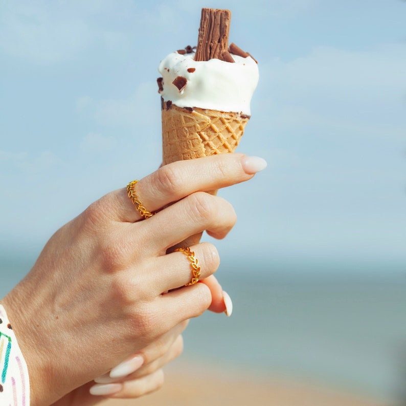May include: A hand wearing two gold rings holds a waffle cone ice cream with chocolate pieces and a chocolate bar on top.