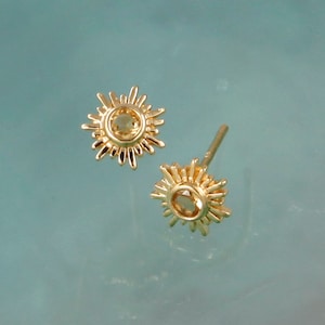 May include: A pair of gold stud earrings shaped like sunbursts, each featuring a small, round, light-colored gemstone set in the center. The earrings have radiating metal rays extending from the central stone, and one earring shows a post backing.