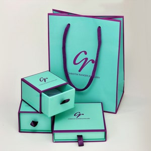 May include: A set of three turquoise gift boxes with purple trim and a drawer. The boxes have the logo "cr" for "Christin Ranger Jewellery" printed on them. The largest box is a shopping bag with purple handles.