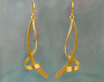 Puede incluir: Un par de pendientes colgantes de tono dorado con un diseño texturizado similar a una cinta que se retuerce en forma de espiral. Cada pendiente cuelga de un gancho sencillo y presenta un acabado mate que refleja la luz en la superficie curva.