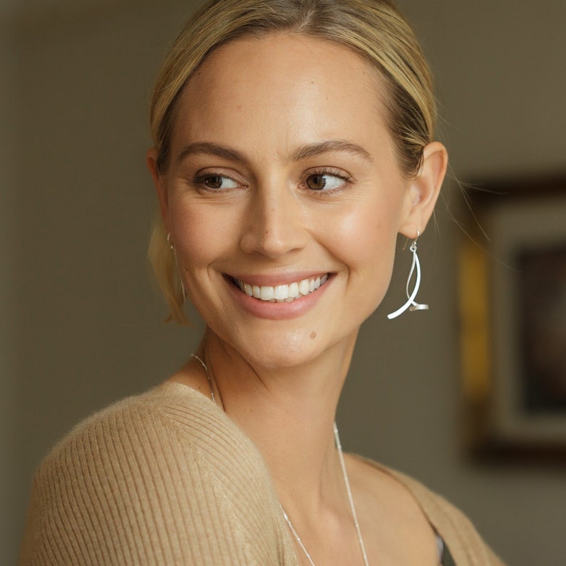 May include: A woman with blonde hair is wearing a silver pair of dangle earrings. The earrings are made of thin, curved metal and have a modern design.