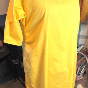 May include: A bright yellow knee-length dress with a square neckline and short sleeves. The garment features a simple, straight-cut silhouette with a smooth, solid-coloured fabric finish, providing a clean and uniform appearance throughout the piece.