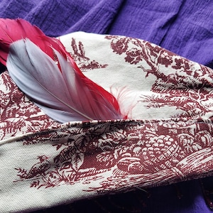 May include: A white fabric hat with a red and white feather attached. The hat has a brown and white toile pattern with a repeating design of trees and houses.