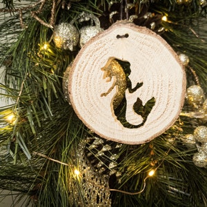 May include: A wooden Christmas ornament shaped like a circle with a mermaid silhouette carved into it. The ornament is hanging on a green pine branch with gold glitter accents.