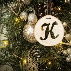 May include: A wooden Christmas bauble with the letter "K" cut out in the centre. The bauble is hanging on a green pine branch with gold glitter baubles and pine cones.