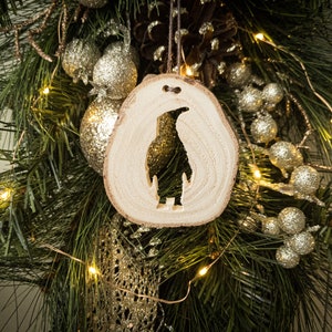 Könnte beinhalten: Ein hölzerner Pinguin-Anhänger mit einem Silhouette-Design, der an einem Weihnachtsbaum mit goldenen und silbernen Ornamenten und Lichtern hängt.