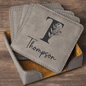 May include: Set of gray leather coasters with a holder. The coasters feature a floral design with the letter T and the name Thompson.