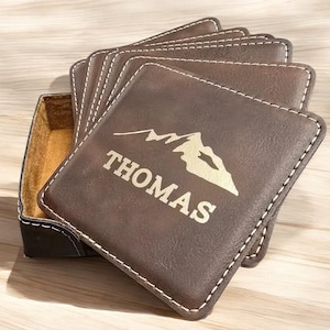 May include: Set of six brown leather coasters with a stitched edge and a gold mountain design. The coasters are in a brown leather holder and the top coaster is personalized with the name "THOMAS".
