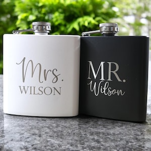 May include: Two stainless steel hip flasks, one white and one black, with the words "Mrs. Wilson" and "MR. Wilson" engraved on them.