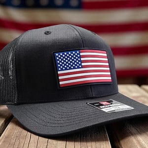 May include: A black trucker hat with a mesh back and a curved brim. The front of the hat features a rectangular patch with the American flag. The hat is sitting on a wooden surface.