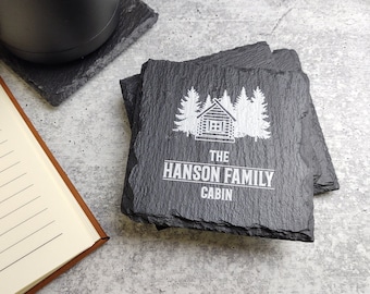 May include: Set of four black slate coasters with a white design of a cabin in the woods, trees, and the text "The Hanson Family Cabin".