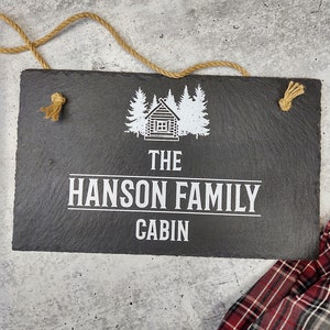 Rustic charcoal black slate sign with a customizable UV ink design featuring a log cabin illustration between pine trees and the text 'THE HANSON FAMILY CABIN' in white. The sign is hung with natural rope on a speckled surface, with a red and gray plaid fabric nearby, evoking a cozy cabin aesthetic.
