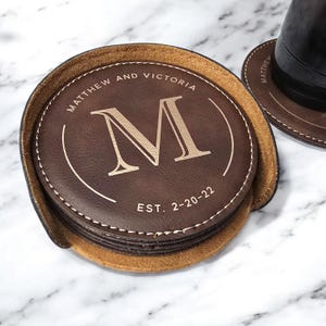 K&ouml;nnte beinhalten: Set aus braunen Lederuntersetzern mit einem gen&auml;hten Rand. Der oberste Untersetzer zeigt den Buchstaben "M" in Gold, den Text "Matthew and Victoria" und "EST. 2-20-22".