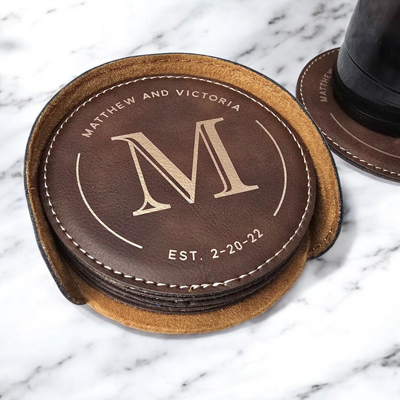 K&ouml;nnte beinhalten: Set aus braunen Lederuntersetzern mit einem gen&auml;hten Rand. Der oberste Untersetzer zeigt den Buchstaben "M" in Gold, den Text "Matthew and Victoria" und "EST. 2-20-22".