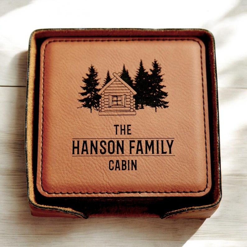 May include: Brown leather coasters with a cabin and trees design. The text "The Hanson Family Cabin" is engraved on the coasters.