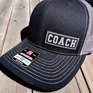 May include: Black trucker hat with a gray mesh back and white stitching. The front panel features the word "COACH" in white block letters within a white rectangular border. The hat has an adjustable strap.
