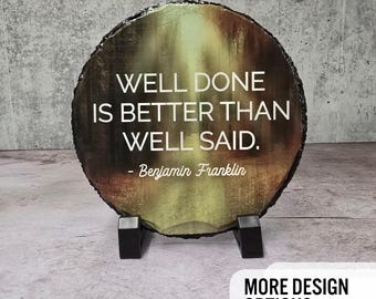 May include: A round slate coaster featuring a forest scene and the quote "Well done is better than well said. - Benjamin Franklin".
