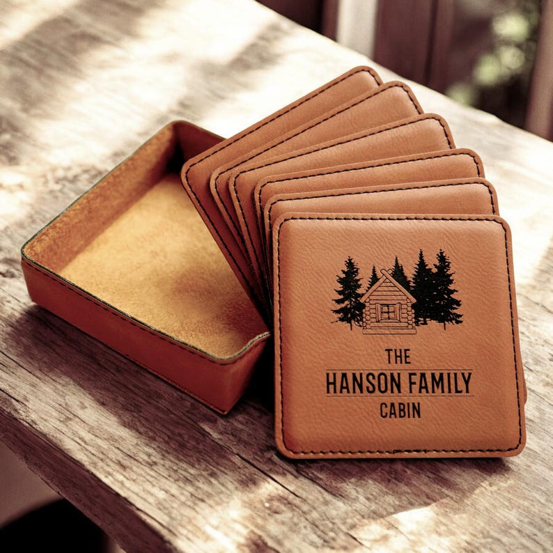 May include: Set of six brown leather coasters with a cabin and trees design. The coasters are in a brown leather holder. The text on the coasters reads "The Hanson Family Cabin".