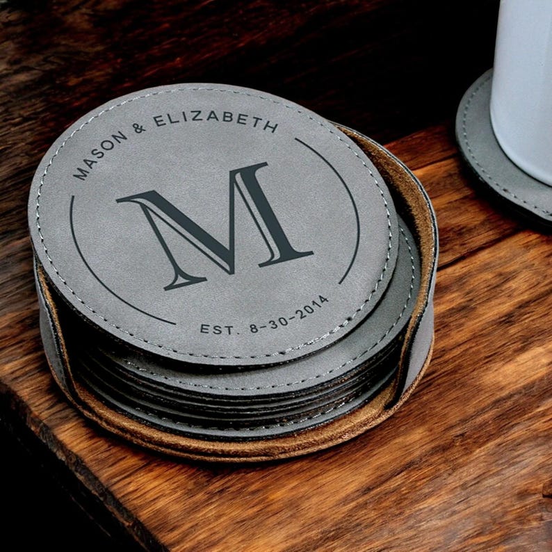 K&ouml;nnte beinhalten: Satz mit sechs grauen Lederuntersetzern mit einer braunen Lederbasis. Der oberste Untersetzer zeigt ein Monogramm "M" mit dem Text "MASON & ELIZABETH EST. 8-30-2014".