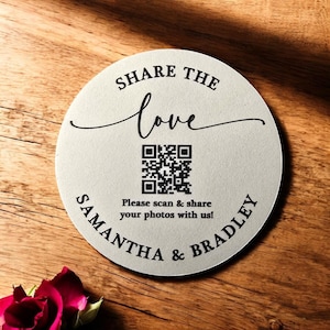 May include: A round, white, wooden sign with black text that reads "Share the love" in a cursive font. A black and white QR code is below the text, with the text "Please scan & share your photos with us!" below the QR code. The text "Samantha & Bradley" is written in a circular arc around the bottom of the sign.