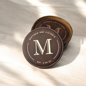 K&ouml;nnte beinhalten: Zwei braune Lederuntersetzer mit einem gen&auml;hten Rand. Der obere Untersetzer tr&auml;gt den Text "Matthew and Victoria" und den Buchstaben "M" in einem Kreis mit dem Text "Est. 2-20-22".
